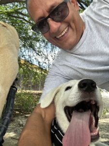 Man wearing sunglasses smiling with a white dog outdoors under leafy trees; they’re close together in a selfie-like scene.