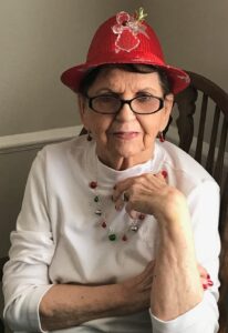 Elderly woman wearing a red sequin hat, glasses, and holiday jewelry sits at a wooden chair indoors.