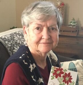 Close-up of an elderly woman with short gray hair, wearing a red top and dark knit vest, holding a holiday card with red poinsettias.