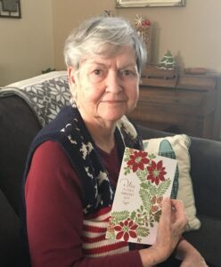 An elderly woman with short white hair sits on a sofa, holding a Christmas card with red poinsettias and green leaves.