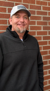 Smiling man with a light beard wearing a white cap and black pullover stands against a red brick wall outside.