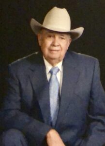Older man wearing a dark suit and light blue tie, pale cowboy hat, seated against a dark background.