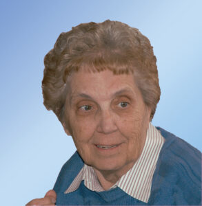 Portrait of an elderly woman with short gray hair wearing a blue sweater and striped collared shirt, against a light blue gradient background