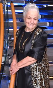 Smiling elderly woman in a black outfit with a leopard-print jacket stands on a glass-and-steel staircase with orange handrails in a modern atrium.