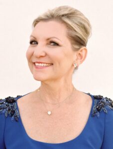 Smiling older woman with short blonde hair, wearing a blue beaded dress and gold jewelry (necklace and earrings).