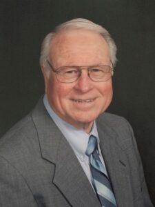 Older man with gray hair and glasses wearing a gray suit and striped blue tie, smiling against a dark background.
