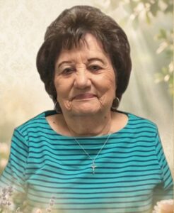 Smiling elderly woman with short dark hair wearing a turquoise striped top and a silver cross necklace.