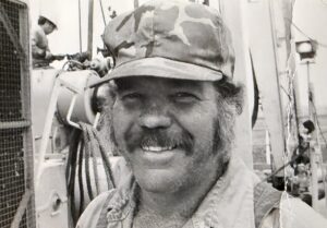 Close-up of a smiling man wearing a camouflage cap and light shirt, with scaffolding and equipment blurred in the black-and-white background.