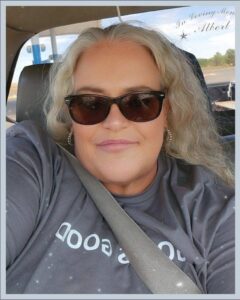 Selfie-style portrait of a woman with wavy blonde hair wearing dark sunglasses, seated in a car with a seat belt across her chest.