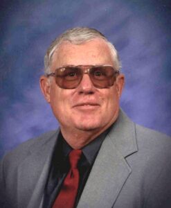 Portrait of an older man wearing brown-tinted sunglasses, a gray suit, black shirt, and red tie in a studio backdrop.