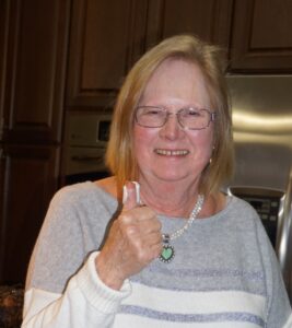 Smiling older woman wearing glasses and a striped sweater gives a thumbs-up in a kitchen.
