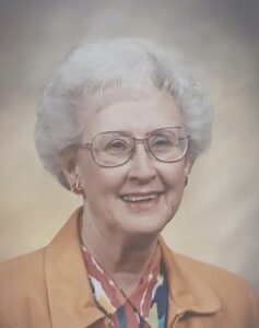 Portrait of an elderly woman with short white hair and glasses, wearing an orange jacket and a multicolored scarf, smiling gently.