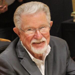 Smiling elderly man with white hair and a beard, wearing glasses and a dark suit.
