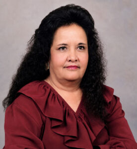 Portrait of a middle-aged woman with long black curly hair, wearing a burgundy ruffled blouse, against a neutral backdrop.