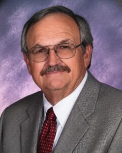 Older gentleman in a gray checkered suit, white shirt, and burgundy tie, posed against a purple studio backdrop for a professional portrait