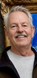 Smiling older man with gray hair and mustache, wearing a light gray shirt and black jacket, ornate gold frame behind him.