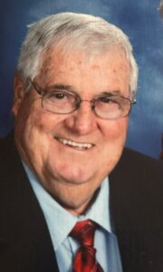 Smiling older man with white hair and glasses in a dark suit and red tie against a blue backdrop.