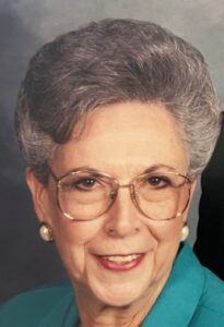 Older woman with short gray hair and round glasses, smiling, wearing a teal blazer and pearl earrings.