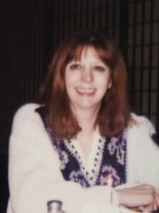 Smiling woman with red hair seated at a table in a dim indoor setting, with condiments (salt shaker, pepper) and a glass in front; handwritten date 11-22-97 visible at the bottom.