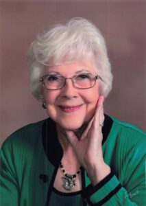 Smiling elderly woman with white hair and glasses, wearing a green cardigan and a black necklace, posed in a neutral studio background.