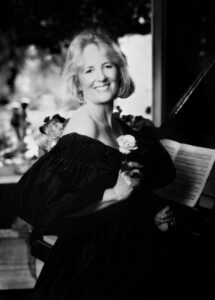 Smiling woman in an off-shoulder dress at a piano with sheet music in her hands.