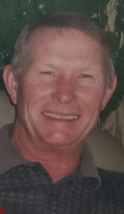 Smiling man facing the camera with short light hair and a collared shirt, outdoors with greenery in the background]