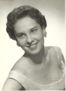Black-and-white studio portrait of a smiling woman with short wavy hair, wearing an off-the-shoulder dress and elegant earrings.