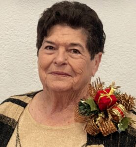 Older woman with short dark hair, wearing a beige knit top and a black-and-gold cardigan, with a festive Christmas corsage on her left shoulder.