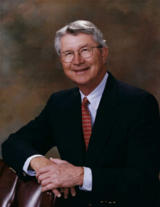 Portrait of an older man with gray hair and glasses, wearing a dark suit and red patterned tie, seated with hands folded on a brown chair.