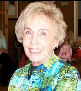 Smiling woman with short blonde hair wearing a bright, colorful patterned shirt at an indoor social event.