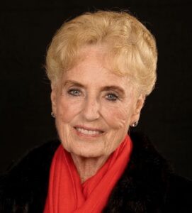 Smiling elderly woman with short blonde hair, wearing a red scarf and black coat against a dark background.