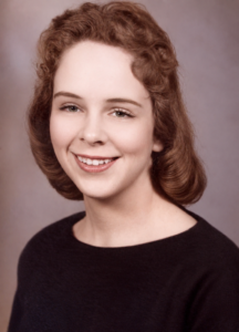 Head-and-shoulders portrait of a smiling woman with short, wavy brown hair wearing a dark sweater in a studio setting.