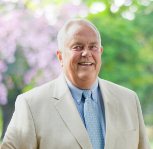 Smiling older man in a light beige suit outdoors with green trees in the background.
