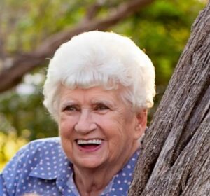Smiling elderly woman with white hair peeking from between tree trunks outdoors.