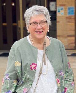 Smiling elderly woman with short gray hair and glasses, wearing a green floral jacket and pearl necklace in front of a building.