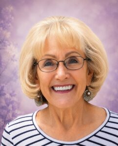 Smiling older woman with short blonde hair and glasses, wearing a striped top and hoop earrings, against a purple background.