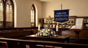 Inside a church sanctuary with wooden pews, stained glass windows, a central altar table holding a floral arrangement and two candles, a cross, and a blue Federated Women's Club banner beside an open Bible on a lectern.