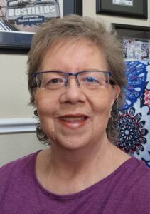 Smiling older woman with short gray hair and blue-framed glasses, wearing a purple top, indoors beside framed decor on the wall.
