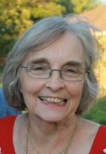 Smiling elderly woman with short gray hair and glasses, wearing a red floral top outdoors.