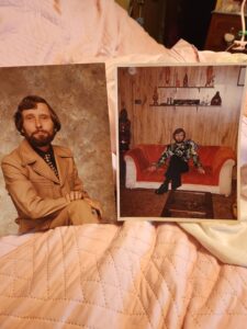 Pair of photos on a bed: left shows a bearded man in a tan suit; right shows a bearded man in a floral shirt sitting on an orange sofa in a wood-paneled room with shelves behind him.