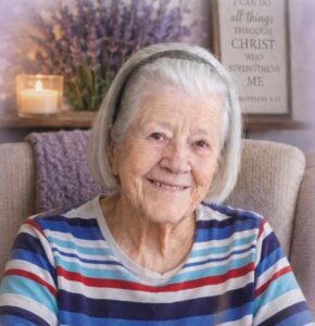 Smiling elderly woman with gray hair in a striped shirt seated on a beige couch, candle and lavender decor in the background.