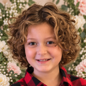 Smiling child with curly brown hair in a red plaid shirt, floral backdrop behind.