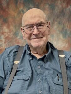 Smiling elderly man with round glasses, bald on top, wearing a blue button-up shirt with suspenders and a mottled background.