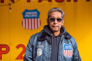 Man with gray hair wearing sunglasses and a blue jacket with patches, standing against a bright yellow backdrop