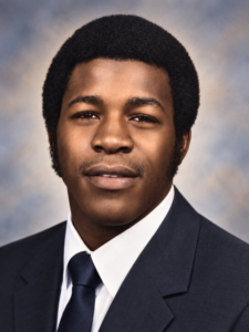 Professional headshot of a young man in a navy suit and tie, facing the camera with a neutral studio backdrop.