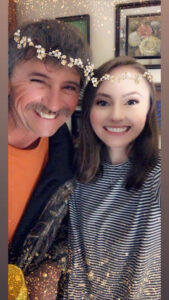 Two smiling people posing for a selfie, a man on the left and a young woman on the right, with a decorative flower crown filter and festive glow effects in the background.
